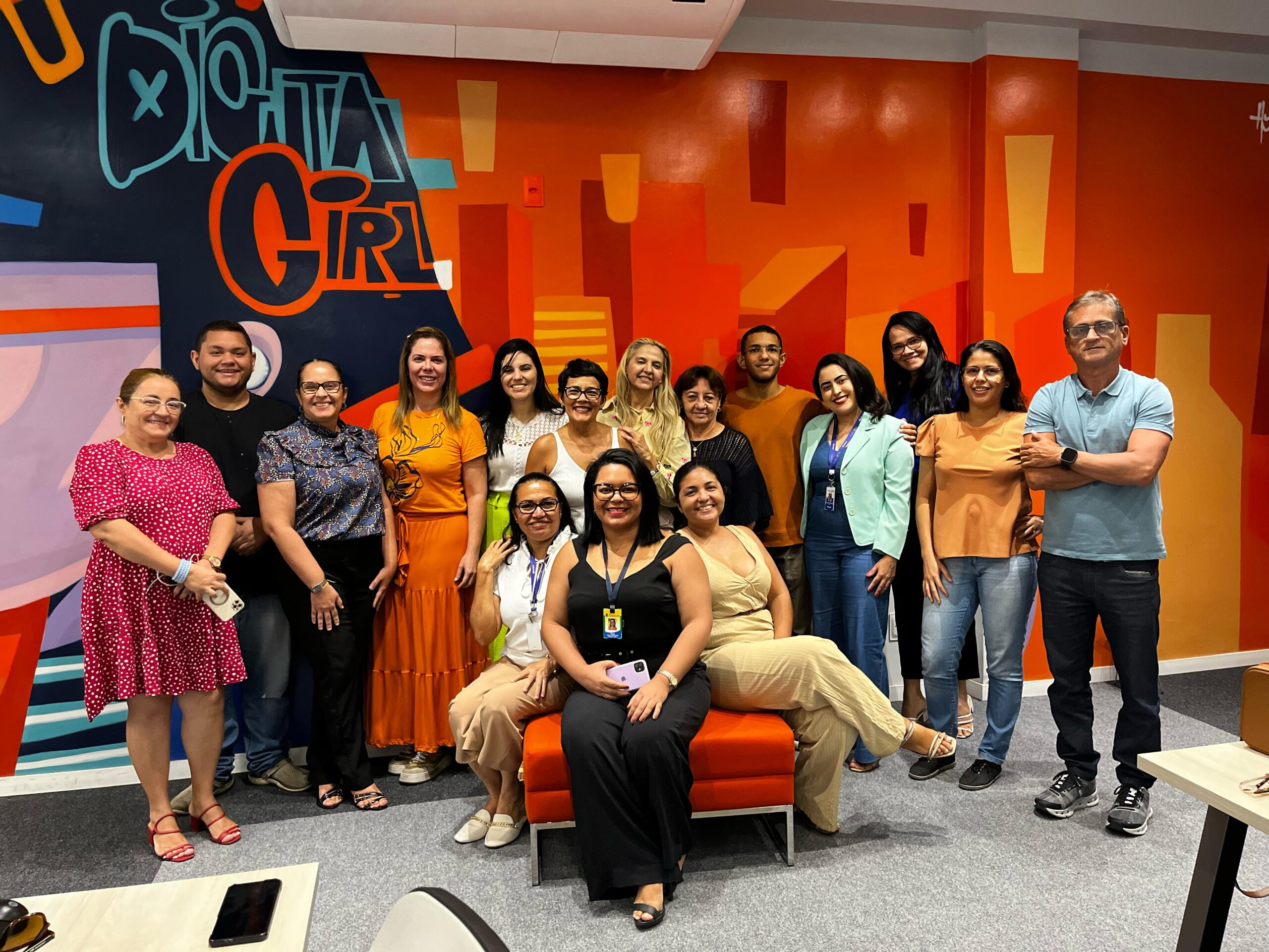 Um grupo de 16 pessoas posa para uma foto em um ambiente interno com decoração moderna e colorida. A parede de fundo apresenta um grafite com as palavras "Digital Girl" em destaque. Os participantes estão sorrindo e vestem roupas casuais e sociais, demonstrando um clima descontraído e acolhedor. Algumas pessoas estão sentadas em um sofá laranja, enquanto outras permanecem em pé ao redor. O ambiente sugere um espaço voltado para tecnologia, inovação ou formação profissional.