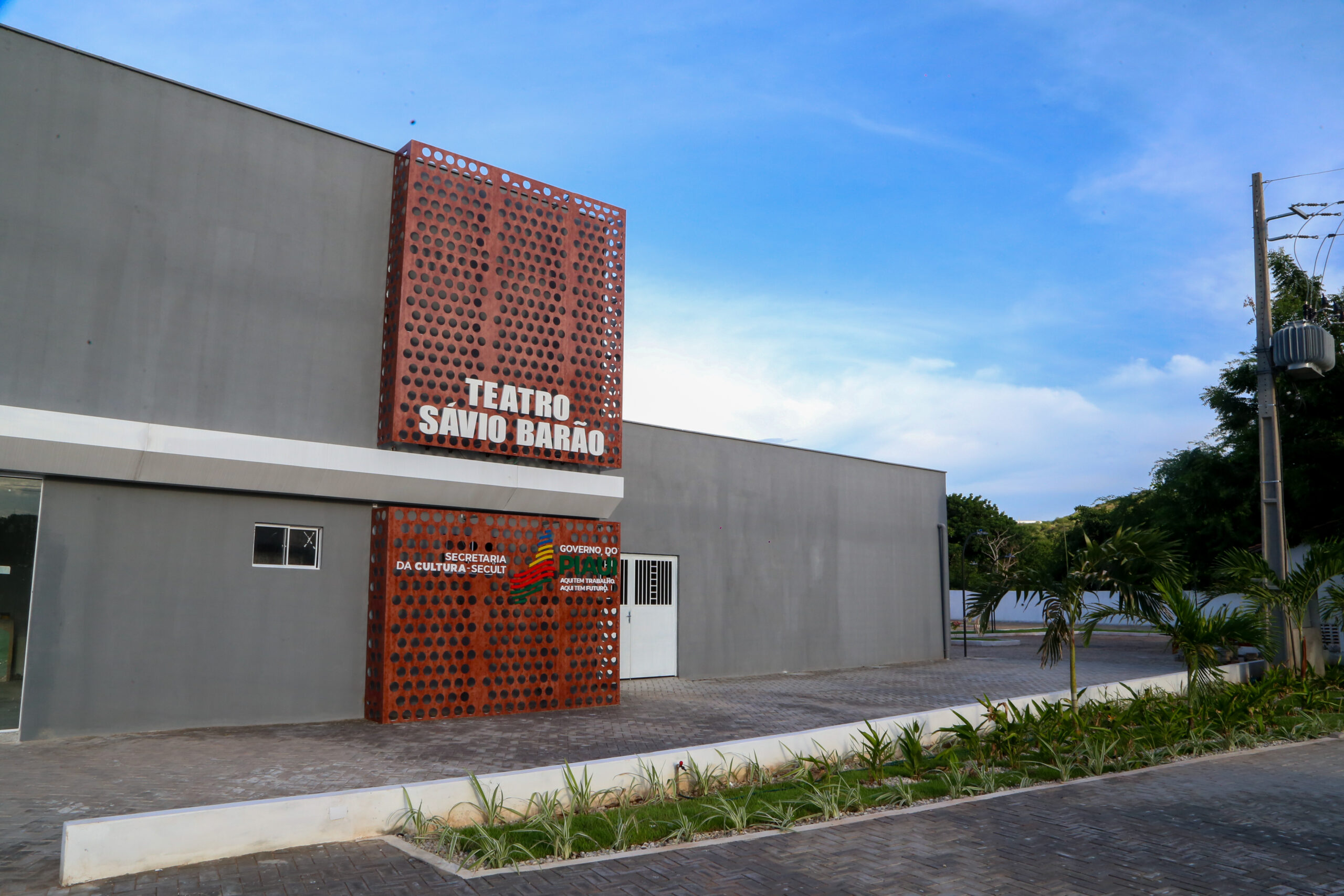 A modern theater building with a distinctive facade, featuring the name "Teatro Savio Barão" prominently displayed. Clear blue sky above.