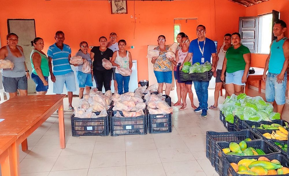 A group of people stands in a room with orange walls, holding bags of food and surrounded by baskets of fresh produce.